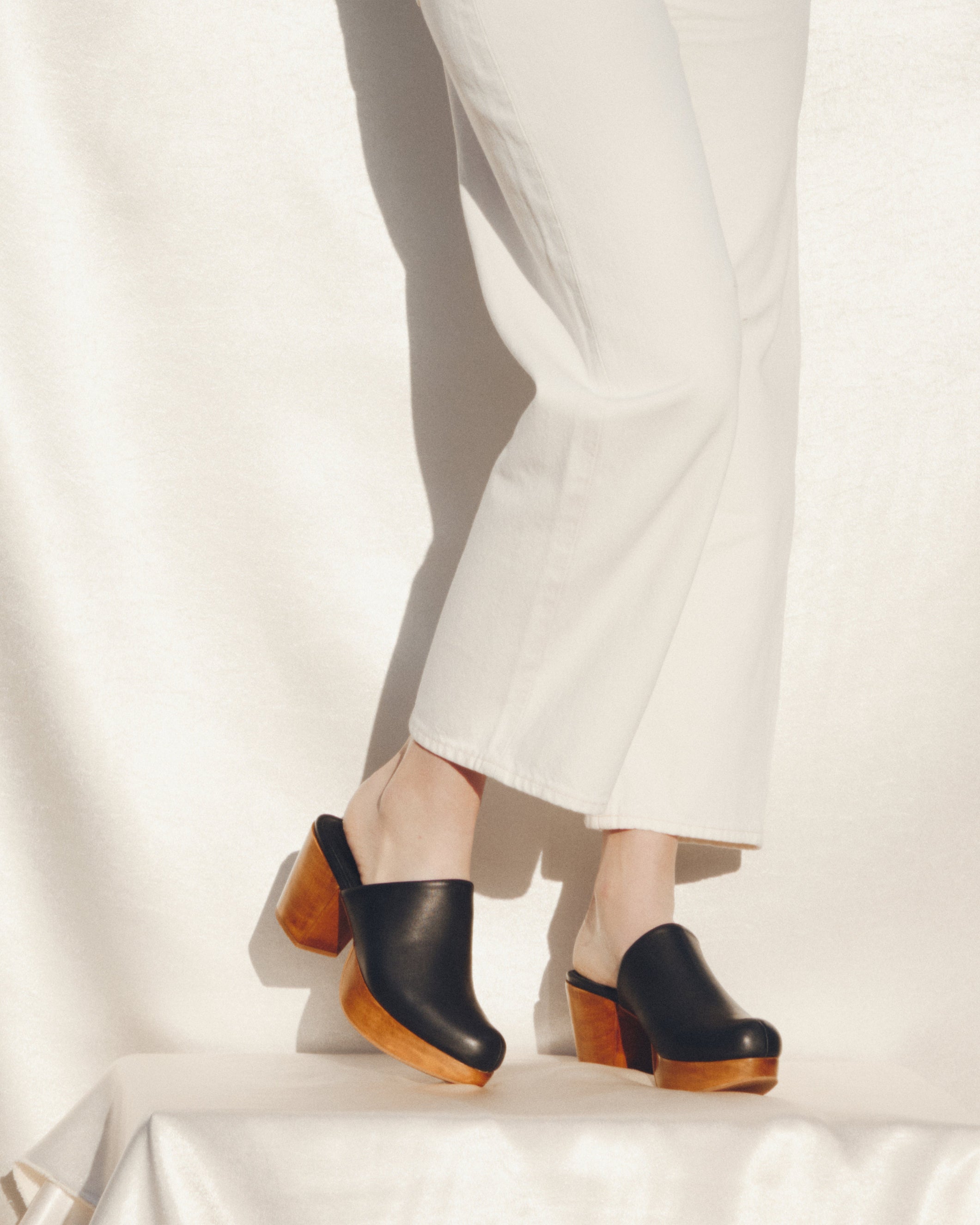 Black clogs with wooden soles worn with white pants on a light background