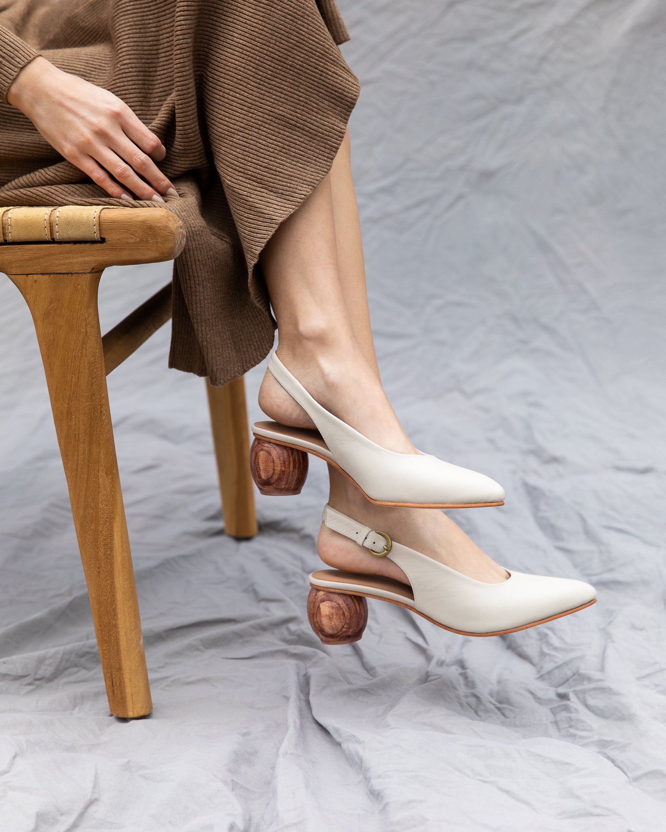 White slingback heels worn by a person sitting on a wooden chair with a neutral background