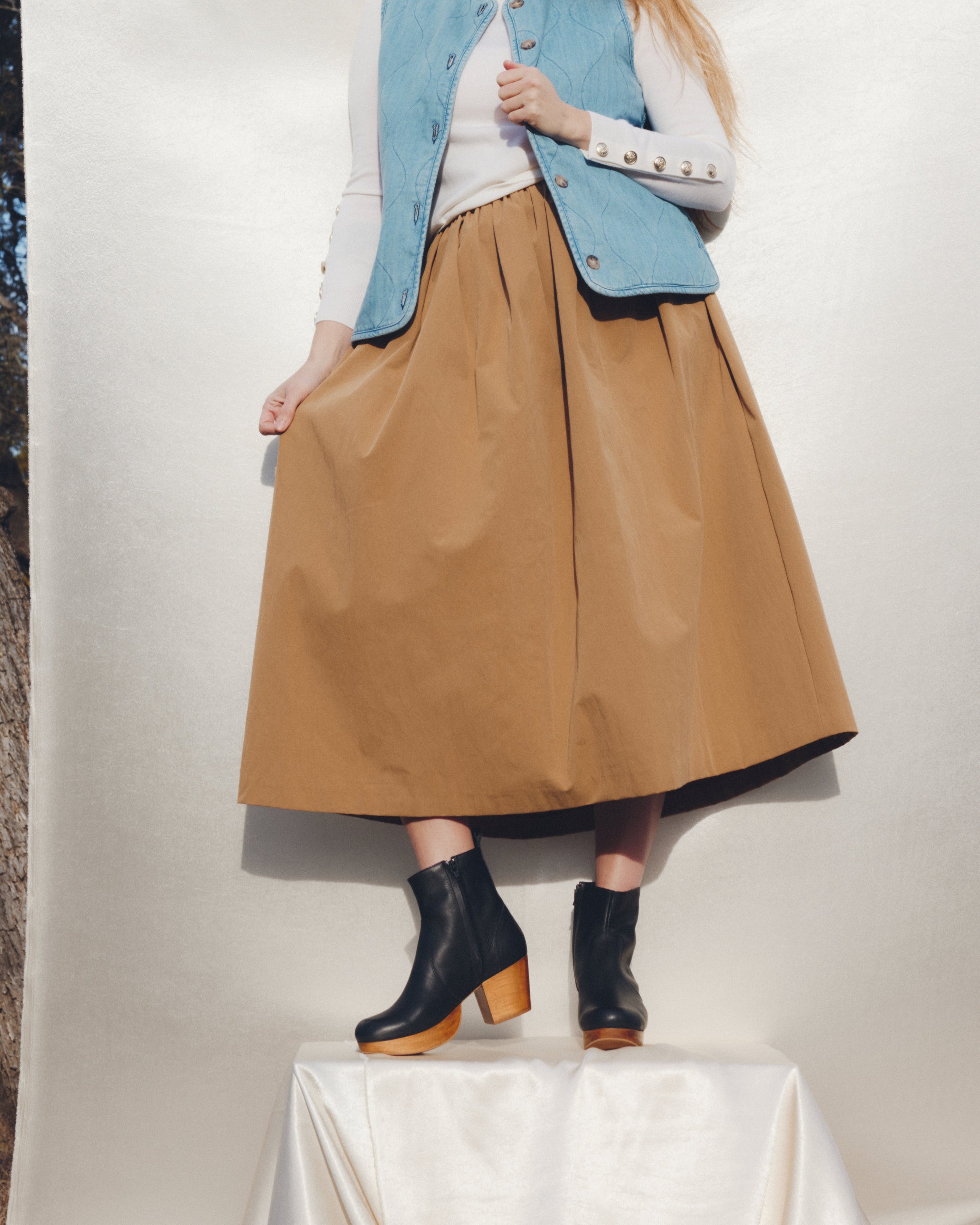 Person wearing a tan skirt, blue vest, white top, and black boots on a white background