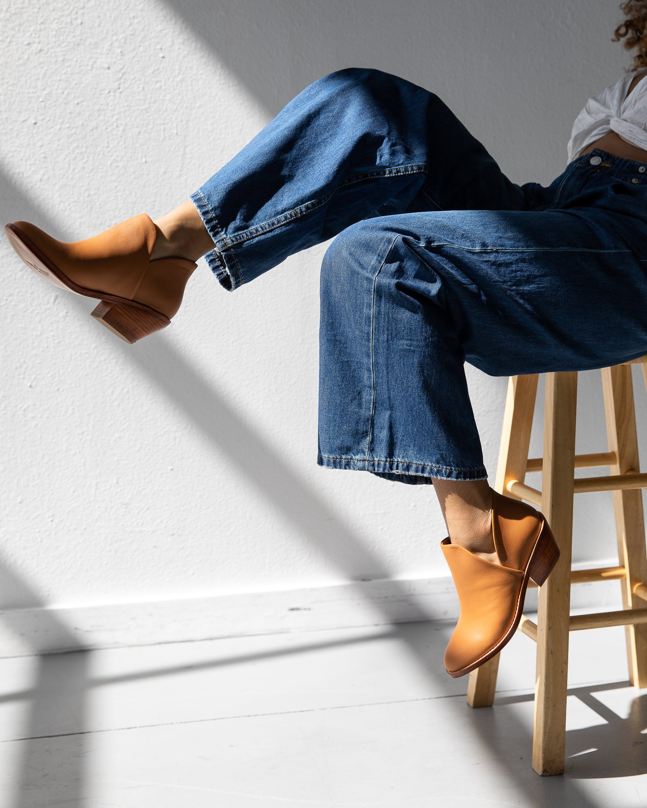 Person wearing blue jeans and brown boots sitting on a wooden stool against a white wall.
