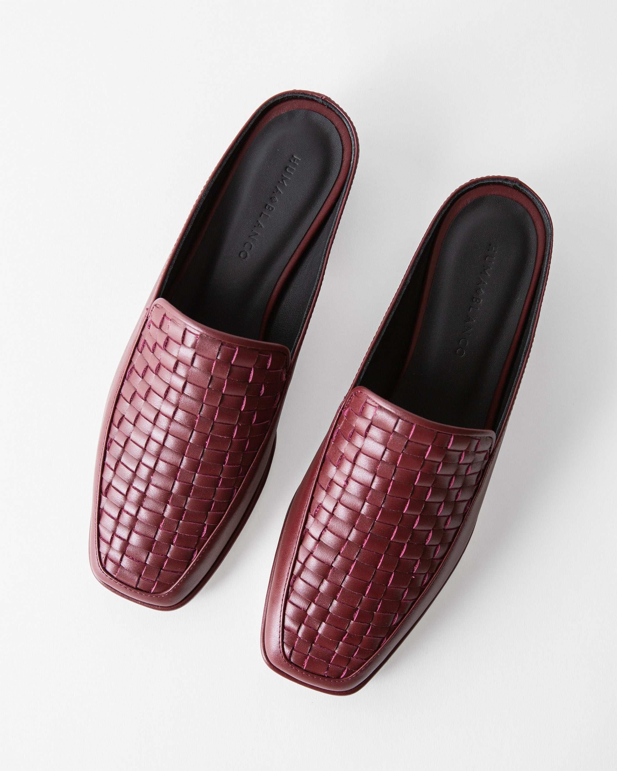 A pair of women's slip-on mule loafers with a woven leather upper and square toes, shown from a top-down angle on a light gray background. The shoes are a deep burgundy/wine color with a black insole.