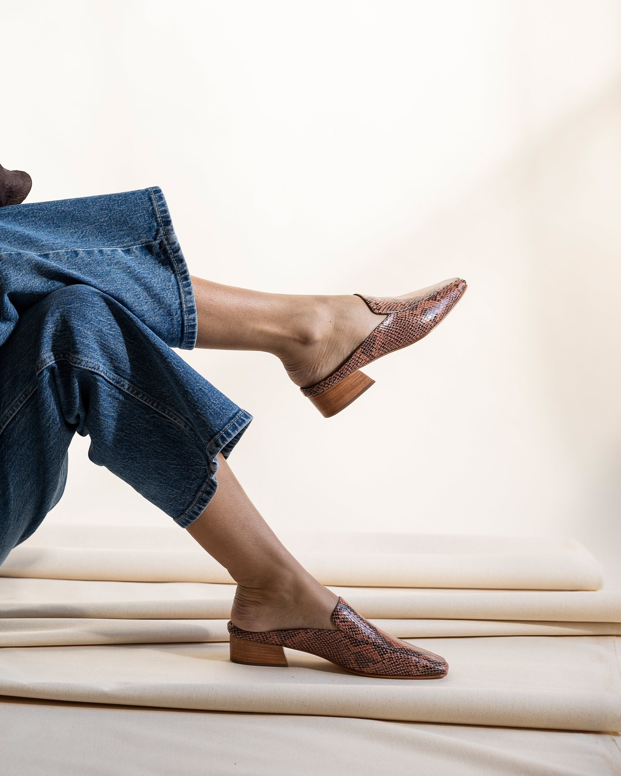 Tan and pink snakeskin loafer mules worn with blue jeans on a light background