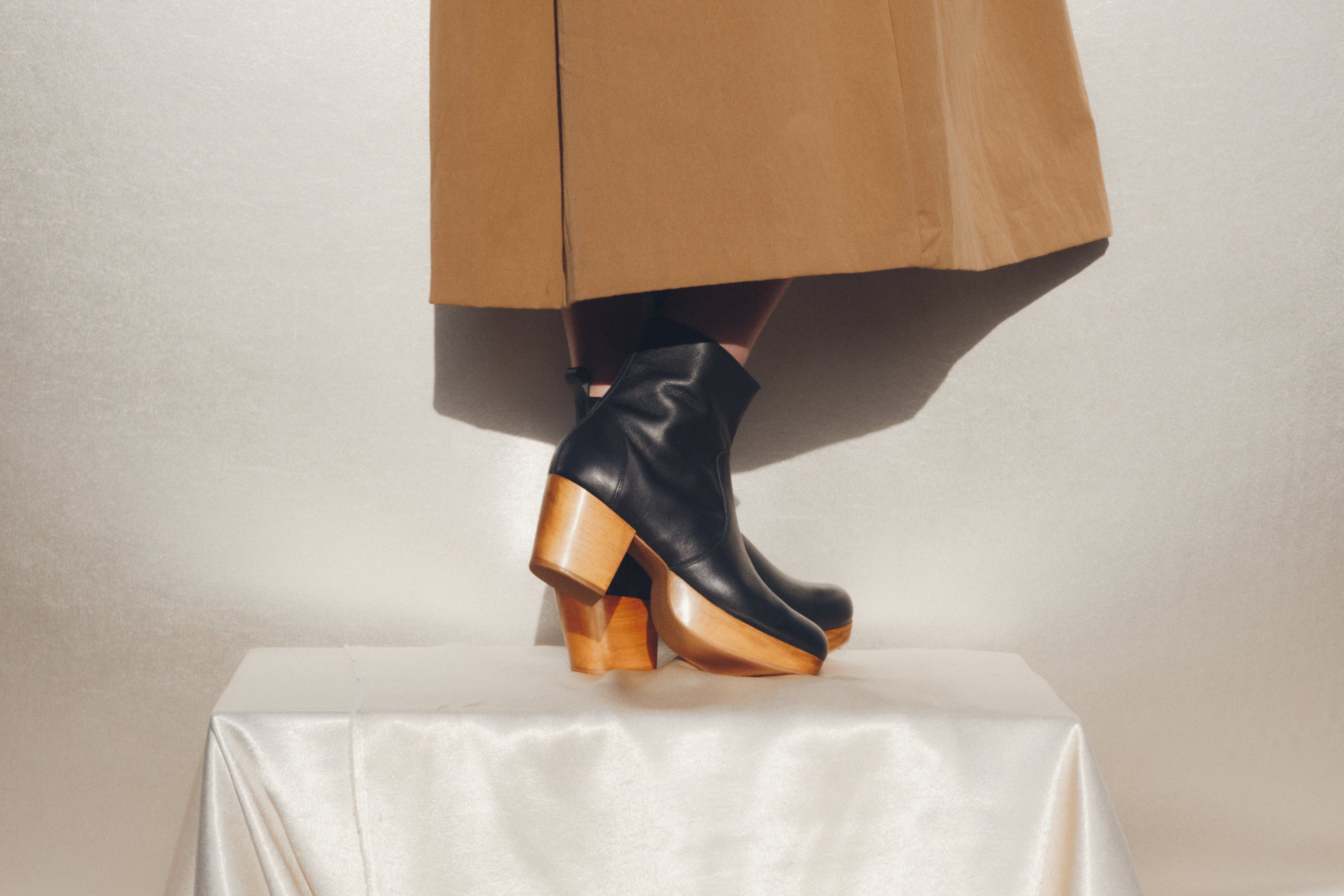 Black ankle boots with wooden soles worn with a tan skirt on a white surface.
