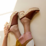 Brown high-heeled shoes with decorative patterns worn by a person sitting on a white surface.