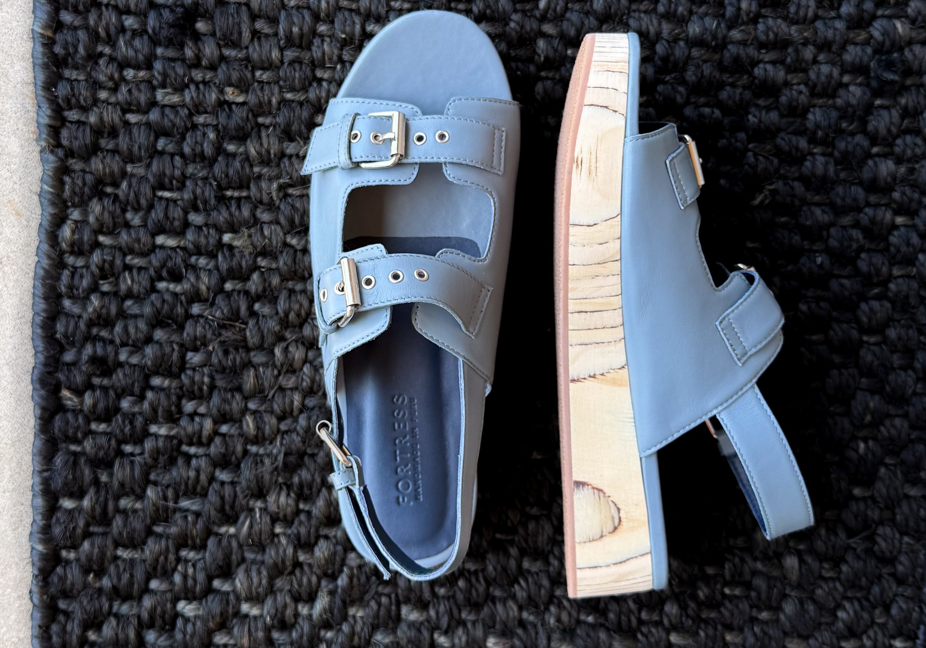 A pair of light blue leather platform sandals with double straps and gold-tone buckles, shown from top and side views on a dark woven rug.