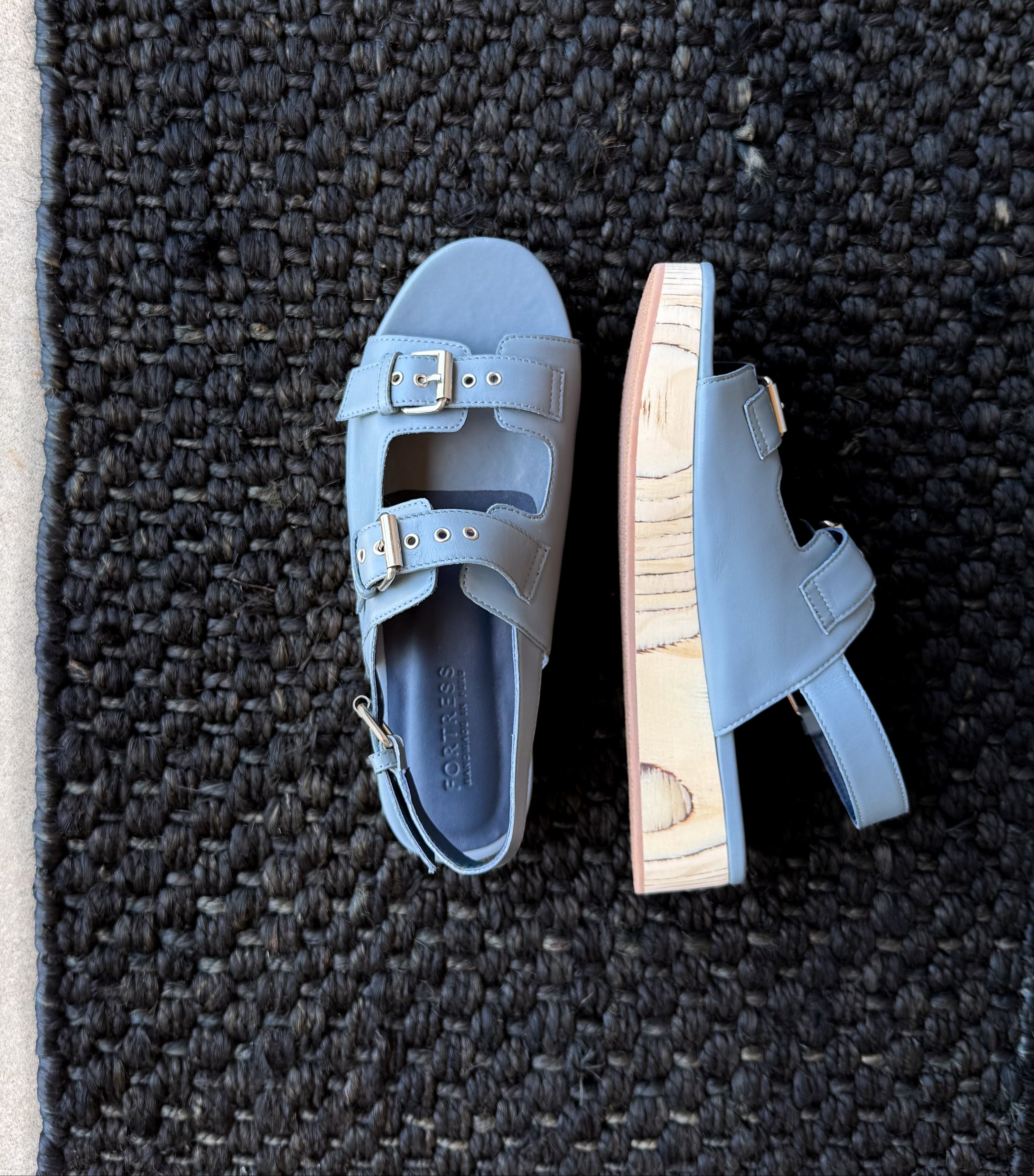 A pair of light blue leather platform sandals with double straps and gold-tone buckles, shown from top and side views on a dark woven rug.