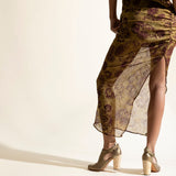 Woman wearing a long, patterned dress, gold heels, with sheer overlay on a beige background