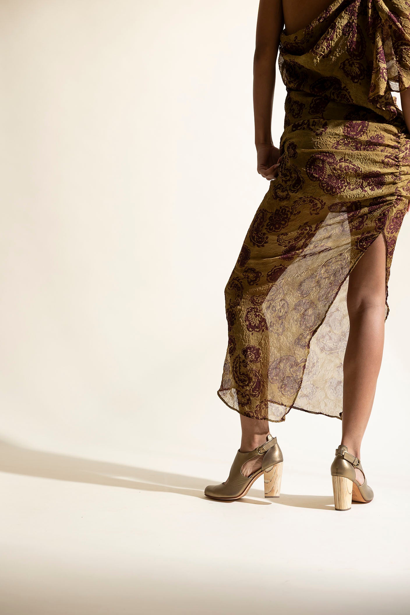 Woman wearing a long, patterned dress, gold heels, with sheer overlay on a beige background