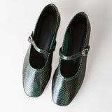 A pair of green snakeskin-patterned Mary Jane shoes with small gold-tone buckle straps, photographed from a top-down angle on a light off-white background.