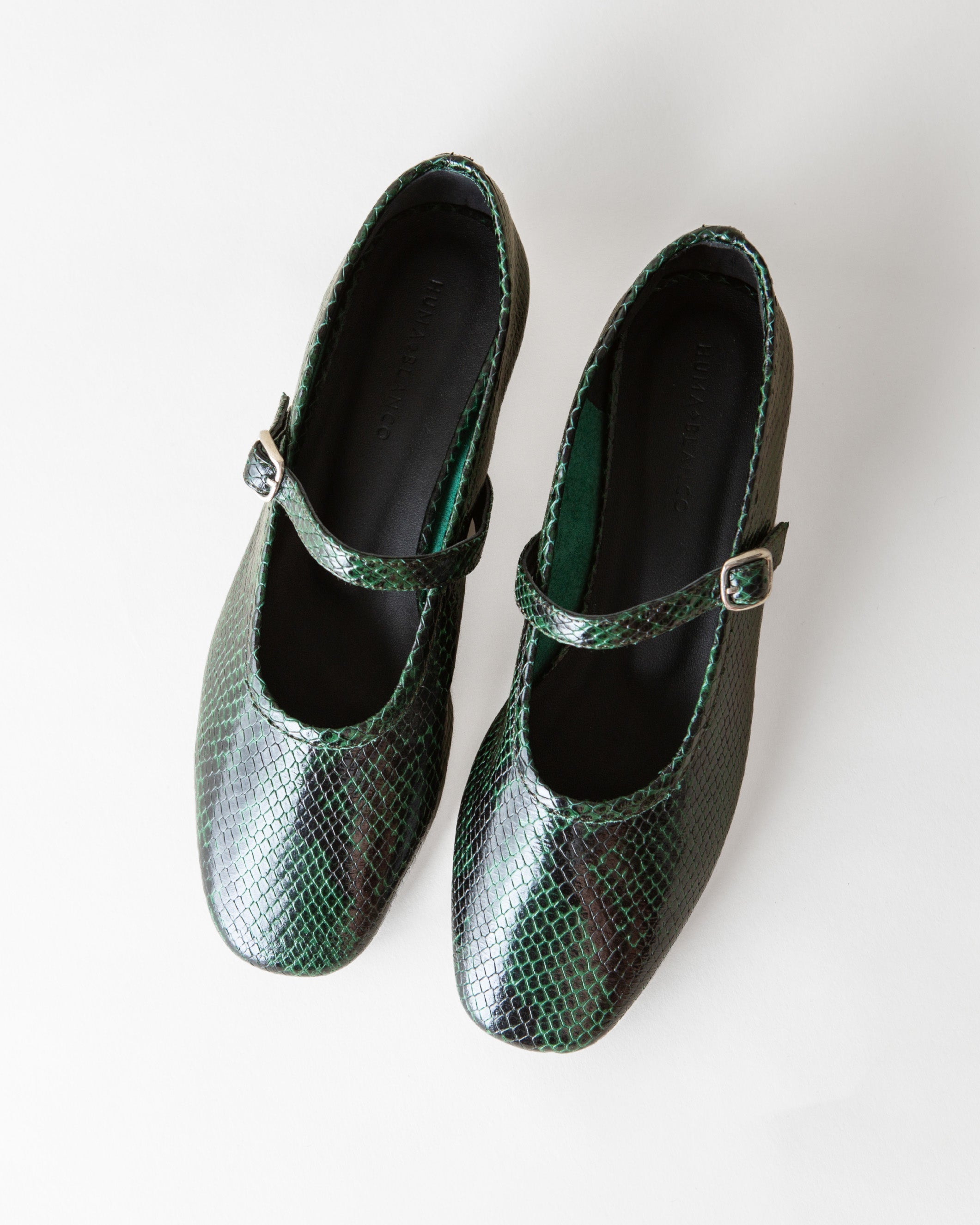 A pair of green snakeskin-patterned Mary Jane shoes with small gold-tone buckle straps, photographed from a top-down angle on a light off-white background.