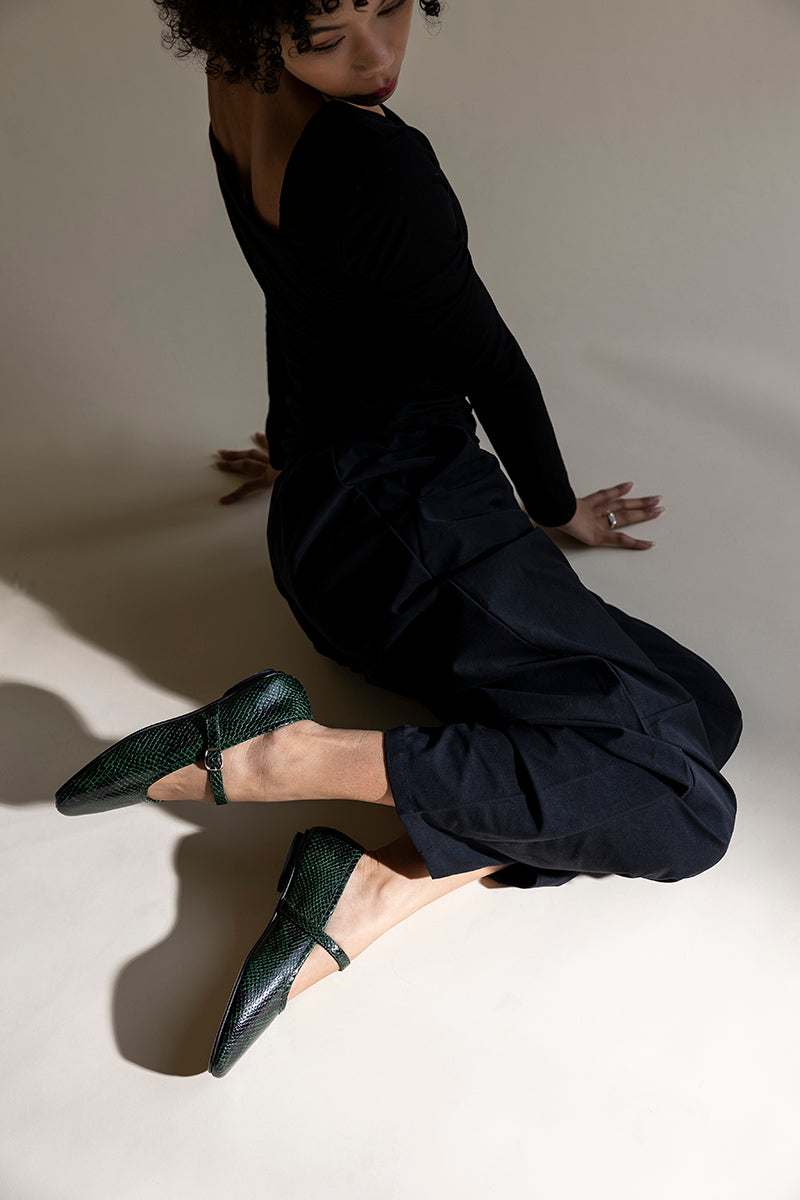 A model in a black long-sleeve top and black pants kneels on a warm beige studio floor, wearing dark green textured Mary Jane flats.