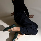 A model in a black long-sleeve top and black pants kneels on a warm beige studio floor, wearing dark green textured Mary Jane flats.
